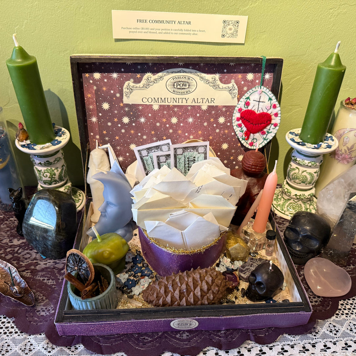 A decorative altar display inside a black tray labeled 'Community Altar' with a sign above reading 'Free Community Altar.' The display includes two tall green candles in ornate holders, folded white paper flowers, crystals, stones, a small plant, and other ritual items arranged on a lace-covered surface against a pale green wall.